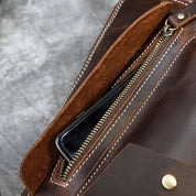 Borsa in pelle marrone con cerniera dorata, cuciture bianche e tasca interna contenente uno smartphone. Accessorio moda elegante e funzionale.