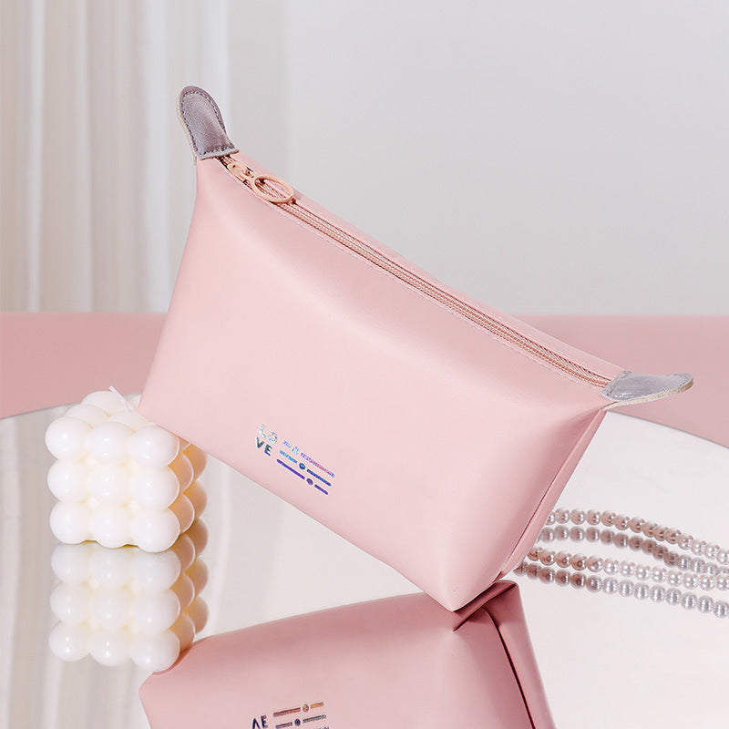 Astuccio rosa in pelle sintetica con cerniera dorata, accanto a candela decorativa bianca e collana di perle. Design elegante e moderno.