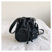 Zaino a secchiello nero in tessuto, con tasca frontale con zip e chiusura a coulisse, su letto bianco. Accessorio moda donna elegante e pratico.