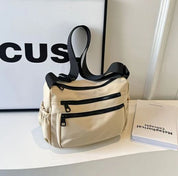 Borsa a tracolla beige con cerniere nere su mensola bianca, accanto a libro aperto. Design moderno, ideale per moda urbana e praticità quotidiana.