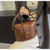 Borsa a tracolla in pelle marrone con chiusura dorata, indossata da una donna in cappotto grigio. Accessorio moda elegante e versatile.