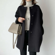 Cappotto nero elegante donna, abbinato a maglione grigio e borsa a tracolla beige. Moda autunnale, stile minimalista, abbigliamento casual chic.