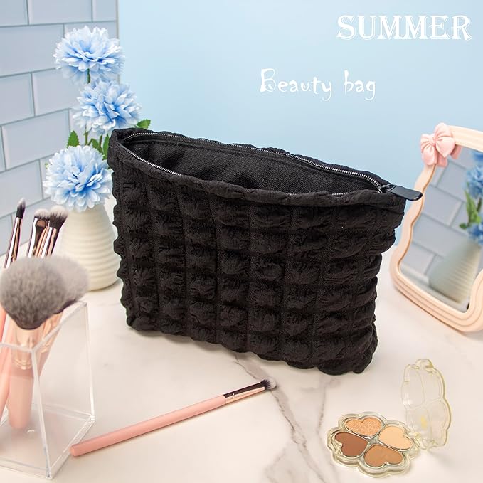 Borsa trucco nera in tessuto trapuntato su tavolo da trucco, circondata da pennelli, fiori blu e specchio. Accessorio essenziale per cosmetici.