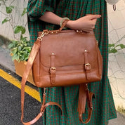 Borsa a tracolla in pelle marrone con fibbie dorate, indossata da una persona in abito a quadri verdi. Accessorio moda elegante e versatile.