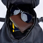 Occhiali da sole neri, portafoglio in pelle marrone e penne in uno zaino nero aperto. Accessori moda, viaggio, organizzazione.