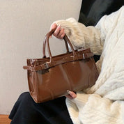 Borsa a mano marrone lucida in pelle, con doppio manico e dettagli a fibbia, indossata da una persona con maglione bianco. Elegante e alla moda.