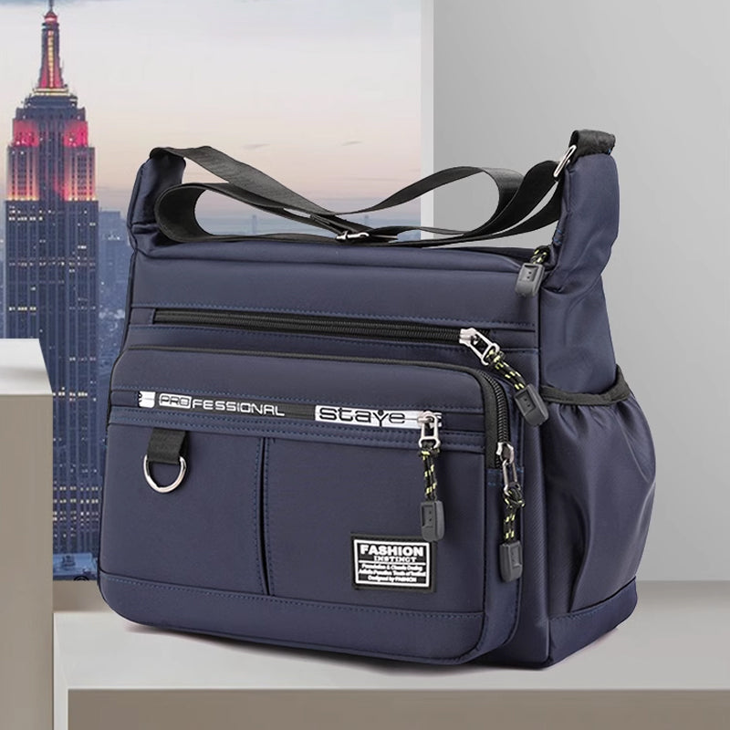 Borsa a tracolla blu scuro con tasche multiple, zip e dettagli in metallo, posizionata su sfondo urbano. Accessorio moda pratico ed elegante.