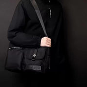 Borsa a tracolla nera unisex, design moderno, con tasche multiple e chiusura a zip, indossata da persona in giacca nera. Accessorio moda versatile.