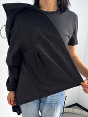 Giacca nera impermeabile da donna, con cappuccio e coulisse, indossata sopra maglietta nera e jeans. Ideale per moda casual e protezione pioggia.