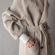 Cappotto beige in lana con cintura, tasche frontali e bottoni, indossato su maglione bianco. Elegante abbigliamento invernale donna.