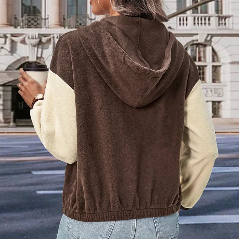 Donna di spalle con felpa con cappuccio bicolore marrone e beige, tiene una tazza di caffè, sfondo urbano. Moda casual autunnale.
