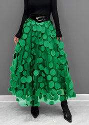 Gonna lunga verde con cerchi decorativi, cintura nera, indossata con top nero a maniche lunghe e stivali a punta. Moda donna elegante e moderna.