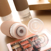 Flacone cosmetico beige con tappo aperto, accanto a palette ombretti. Prodotti di bellezza, trucco, skincare, accessori donna.