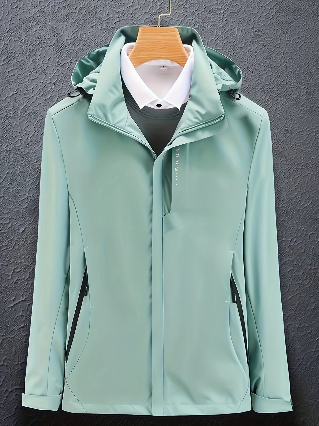 Giacca impermeabile verde menta con cappuccio, appesa su gruccia, su sfondo grigio. Design elegante, ideale per moda uomo e abbigliamento outdoor.
