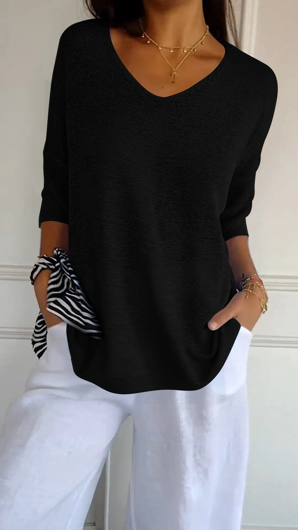 Donna con maglia nera a maniche lunghe, scollo a V, abbinata a pantaloni bianchi. Accessori: collana dorata, bracciali, foulard a righe.