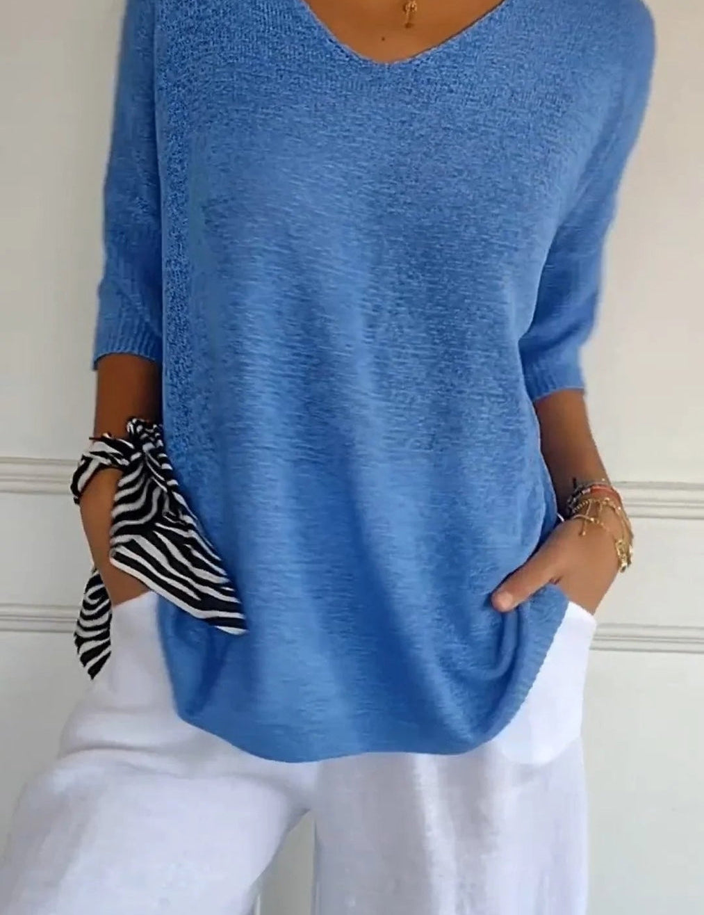 Maglia blu a maniche lunghe con scollo a V, abbinata a pantaloni bianchi. Accessori: bracciali dorati e foulard a righe. Moda casual donna.