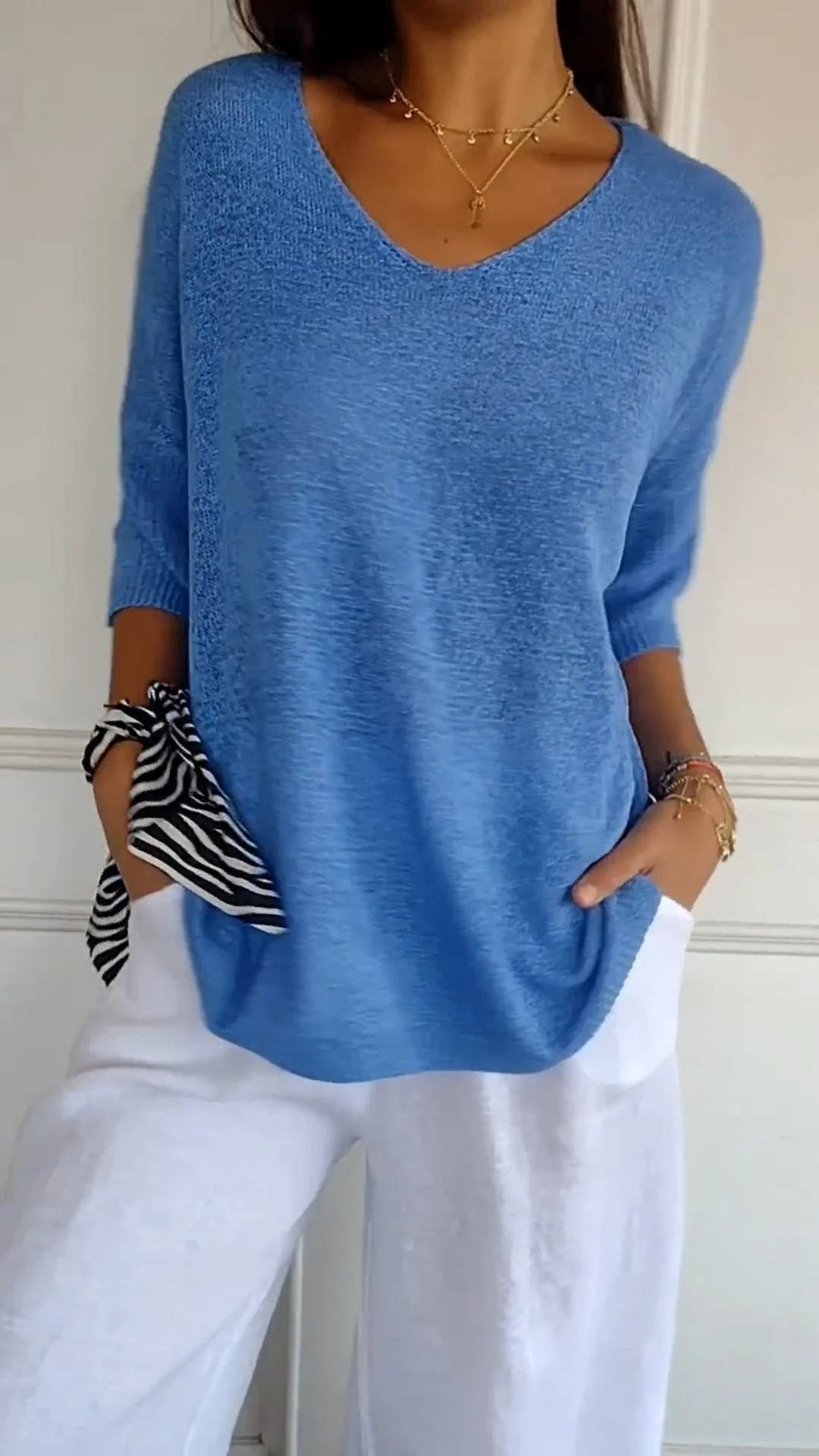 Maglia blu a maniche lunghe con scollo a V, abbinata a pantaloni bianchi. Accessori: bracciali dorati e foulard a righe. Moda casual donna.