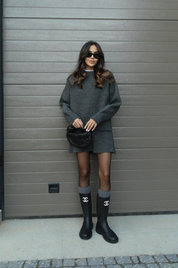 Donna con abito grigio oversize, stivali neri Chanel, borsa a tracolla nera e occhiali da sole, posa davanti a una porta grigia.