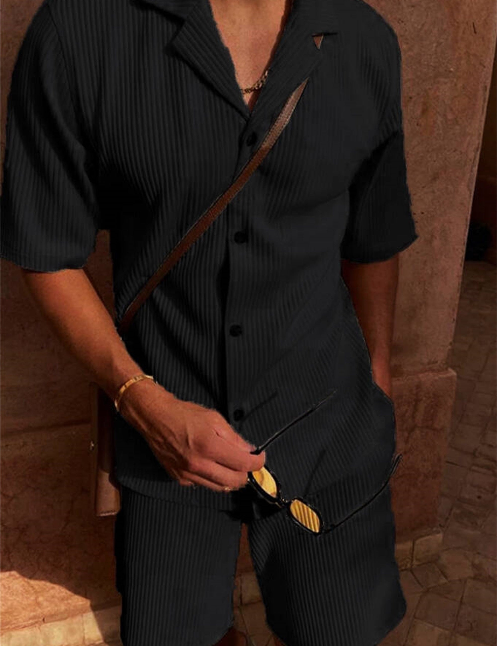 Uomo in completo estivo nero a righe, camicia a maniche corte e pantaloncini coordinati, con occhiali da sole e bracciale dorato.