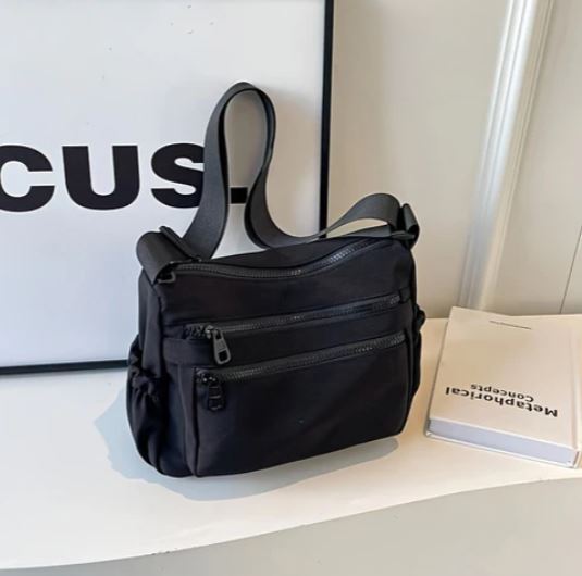 Borsa a tracolla nera su tavolo bianco, con doppia cerniera e tasche multiple. Design moderno, ideale per moda urbana e praticità quotidiana.