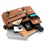 Borsa a tracolla in pelle marrone con tasche multiple, contenente smartphone, chiavi, auricolari, orologio, fotocamera, taccuino, iPad e ombrello.