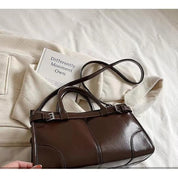 Borsa a mano in pelle marrone su letto bianco, accanto a libro e maglione beige. Elegante accessorio moda donna, stile classico e sofisticato.