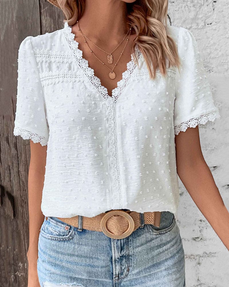 Blusa bianca in pizzo con scollo a V, maniche corte, dettagli traforati, abbinata a jeans chiari e cintura beige. Moda estiva elegante e casual.