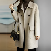 Cappotto donna beige elegante, doppio petto, tasche frontali, abbinato a borsa nera trapuntata. Moda autunnale, stile casual chic.