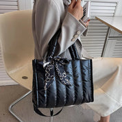 Borsa a tracolla nera trapuntata con catena e fiocco, indossata da una persona seduta su sedia bianca. Elegante accessorio moda femminile.