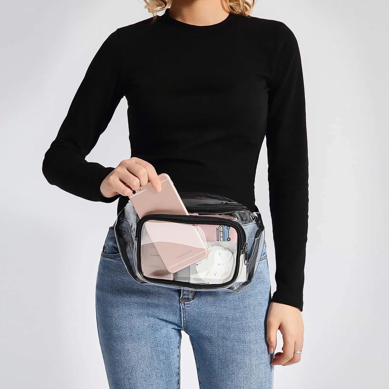 Donna con marsupio trasparente, indossa maglia nera e jeans. Marsupio contiene smartphone e accessori. Moda pratica, accessori trasparenti.