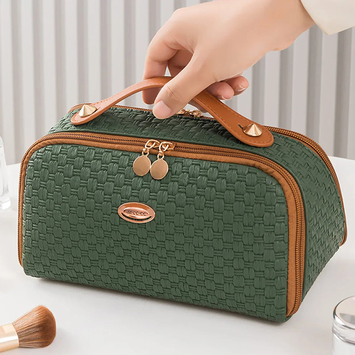 Borsa cosmetica verde intrecciata con manico in pelle marrone, chiusura a zip dorata, ideale per trucco e accessori da viaggio.