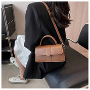 Donna con borsa a tracolla in pelle marrone, seduta su sedia moderna. Look elegante con blazer nero e sneakers bianche. Accessorio moda chic.