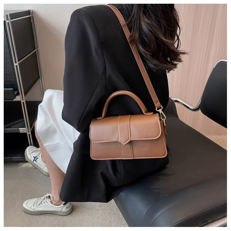 Donna con borsa a tracolla in pelle marrone, seduta su sedia moderna. Look elegante con blazer nero e sneakers bianche. Accessorio moda chic.