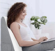 Donna sorridente usa laptop su cuscino ergonomico grigio, pianta verde sullo sfondo. Comfort, lavoro da casa, arredamento moderno.