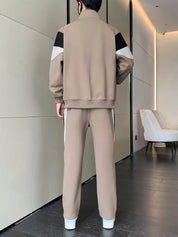 Uomo in tuta sportiva beige con dettagli neri e bianchi, vista posteriore. Abbigliamento casual moderno, pavimento in legno, pareti chiare.