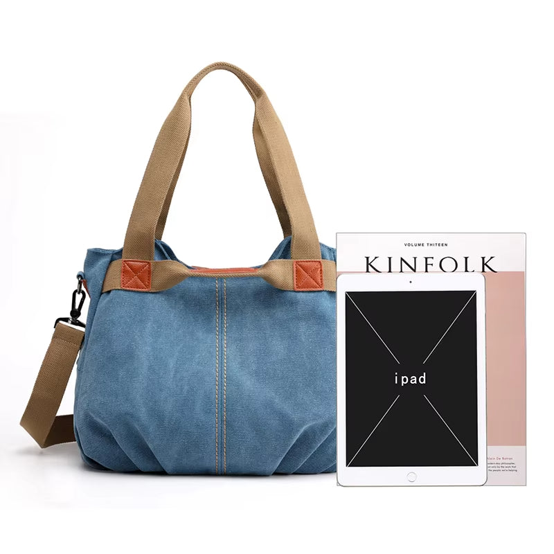 Borsa a tracolla in tela blu con manici marroni, accanto a un iPad e una rivista Kinfolk. Design casual, ideale per uso quotidiano e viaggi.