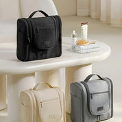 Borse da viaggio appese in nero, beige e grigio su tavolo bianco, con accessori da bagno. Organizzatori cosmetici, eleganti e funzionali.