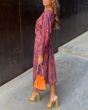 Abito midi viola con motivo floreale arancione, borsa arancione, sandali dorati con tacco alto. Look elegante e colorato per eventi estivi.