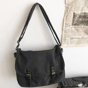 Borsa a tracolla nera in tela con fibbie, appesa a parete bianca. Design casual, ideale per uso quotidiano. Accessorio moda versatile.