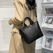 Donna con cappotto beige indossa borsa a tracolla nera in pelle, elegante e moderna, davanti a riviste di moda. Accessorio di tendenza per l'autunno.