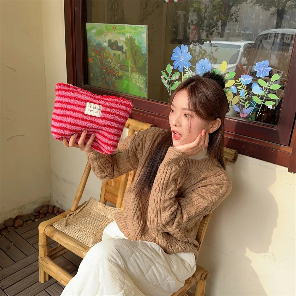Giovane donna con maglione marrone e pantaloni bianchi, seduta su sedia di legno, tiene borsa a righe rosse e bianche. Sfondo con fiori e finestra.