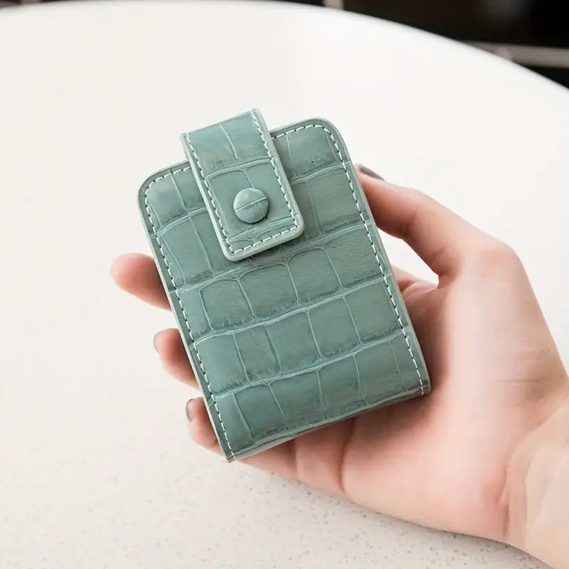 Portafoglio donna in pelle verde con texture coccodrillo, chiusura a bottone, design compatto, elegante e moderno, tenuto in mano su sfondo chiaro.