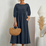 Donna con abito lungo blu scuro, maniche a tre quarti, borsa in paglia, sfondo minimalista. Moda elegante, abbigliamento femminile casual.