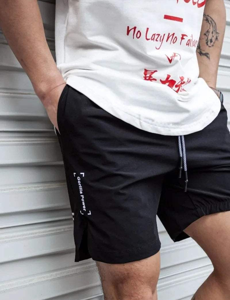 Uomo con pantaloncini sportivi neri e maglietta bianca con scritte rosse, posa casuale. Abbigliamento estivo, moda uomo, stile urbano.