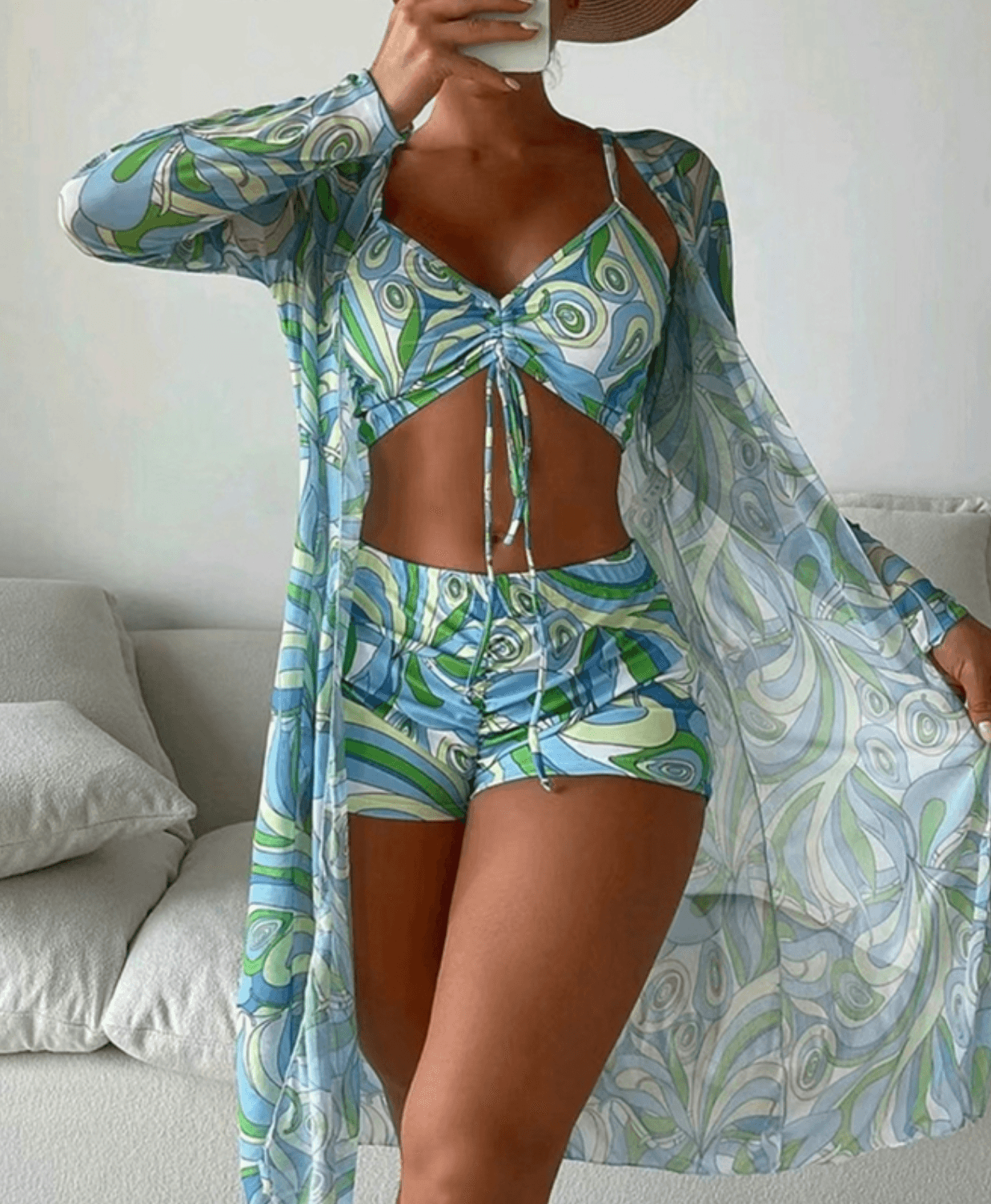 Donna in costume da bagno estivo a fantasia geometrica blu e verde, con copricostume trasparente coordinato, posa in un ambiente interno moderno.