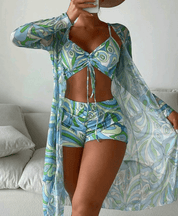 Donna in costume da bagno estivo a fantasia geometrica blu e verde, con copricostume trasparente coordinato, posa in un ambiente interno moderno.