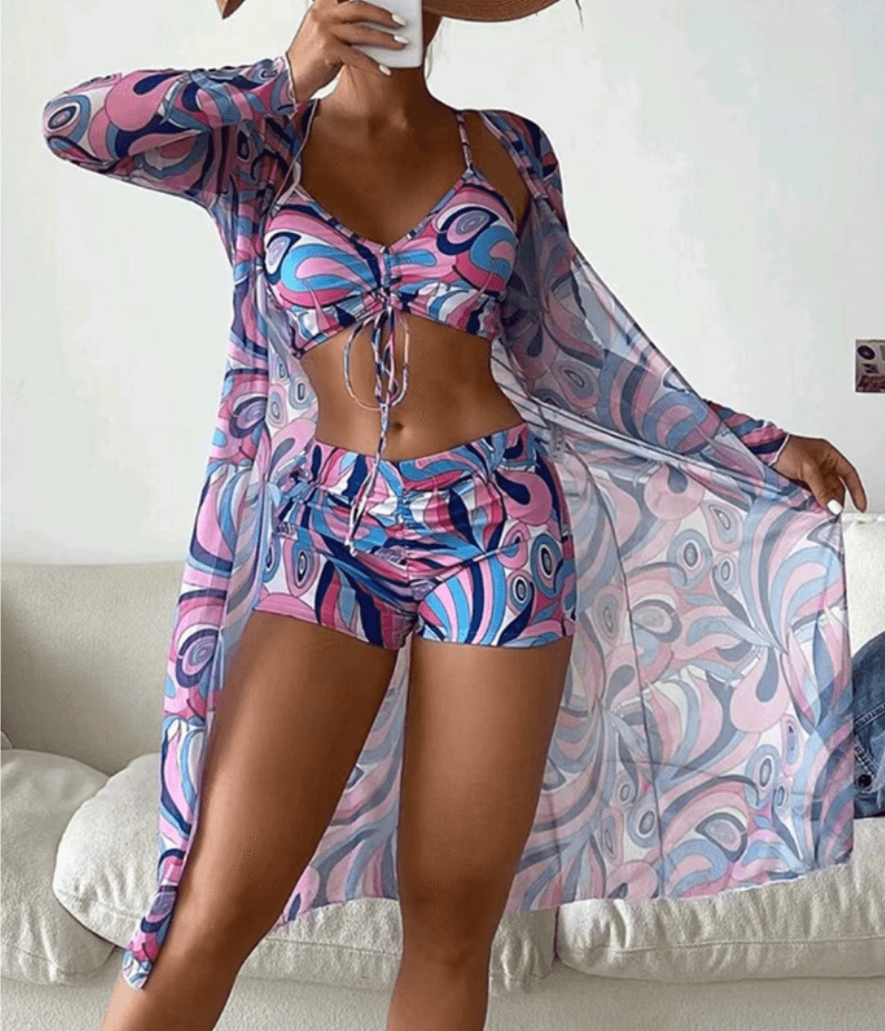 Donna in costume da bagno fantasia psichedelica rosa e blu con copricostume coordinato, posa su divano bianco. Moda estiva, abbigliamento mare.