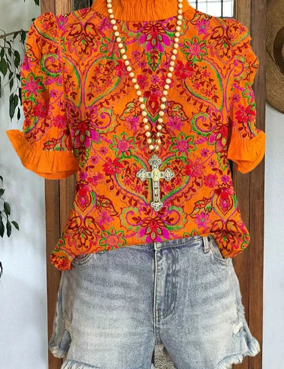 Blusa arancione a fiori con maniche corte e colletto arricciato, abbinata a shorts di jeans sfrangiati. Collana con croce decorativa.