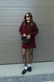 Donna con abito bordeaux, occhiali da sole neri, borsa a mano nera, calze nere e mocassini neri. Moda autunnale elegante e moderna.