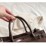 Borsa a mano in pelle marrone lucida con manici, su sfondo di tessuto bianco. Accessorio moda elegante, ideale per shopping e lavoro.